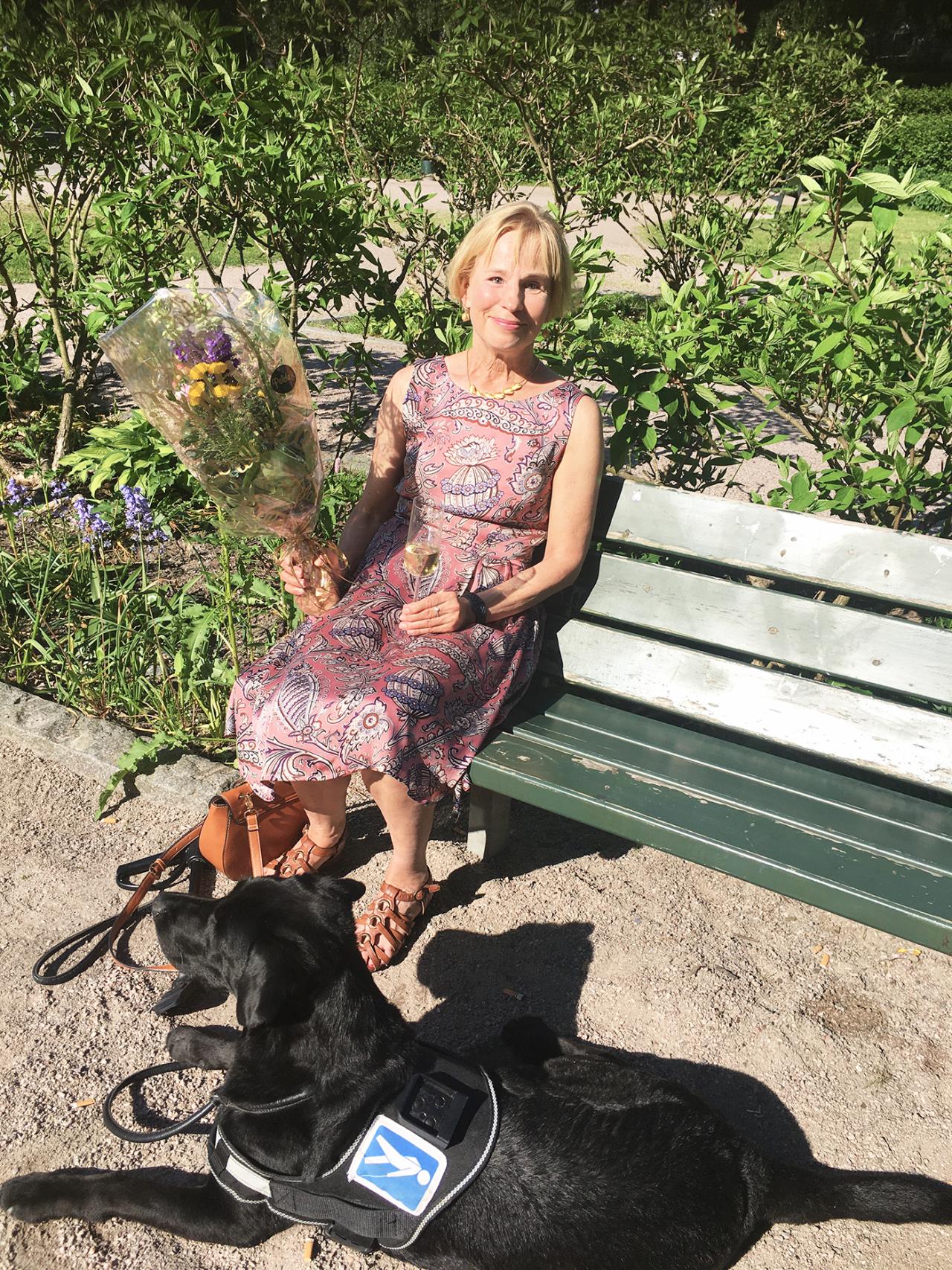 Nainen istuu kesämekossa puistonpenkillä iso kukkakimppu kädessään, opaskoira makaa maassa naisen jaloissa.